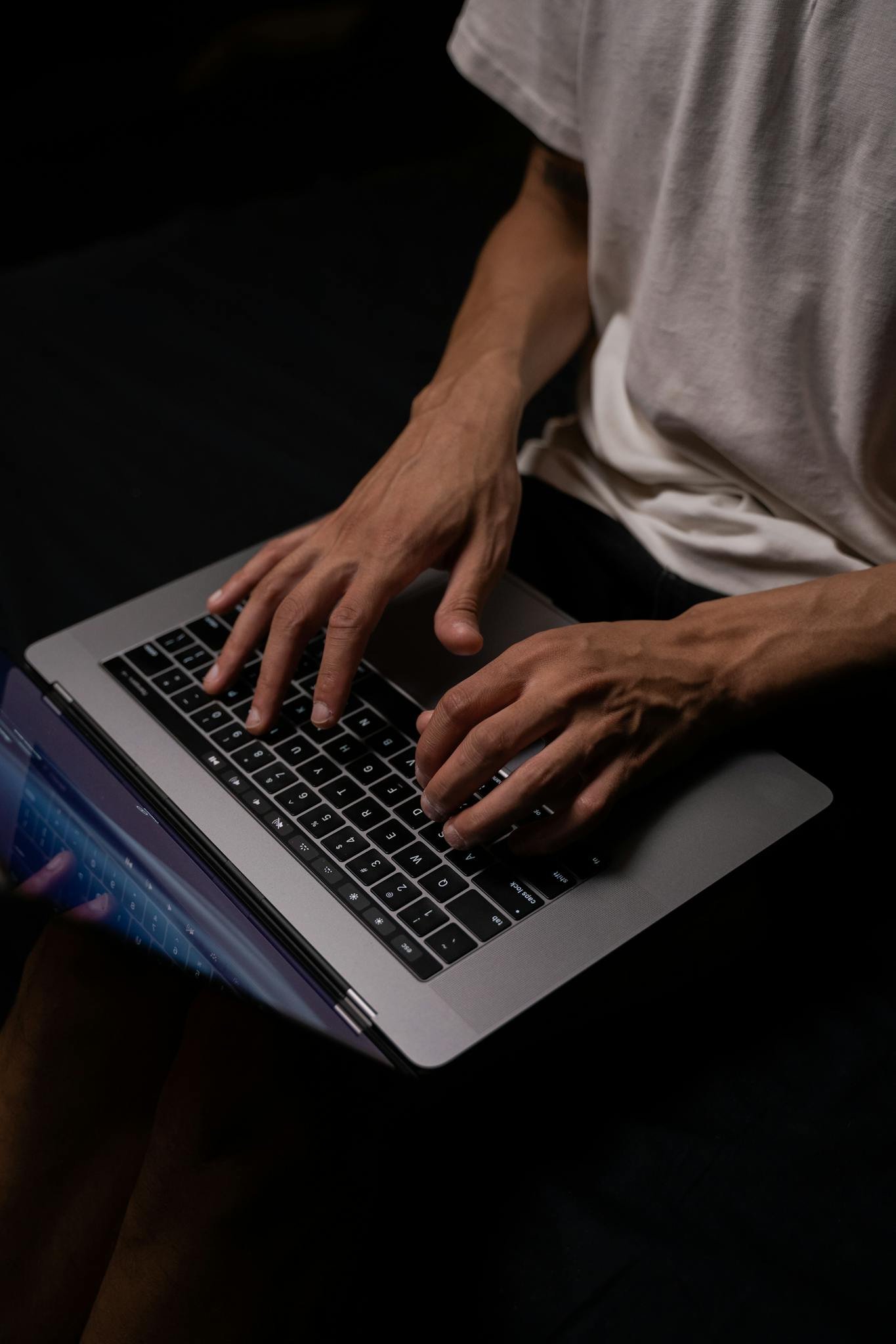 An adult male programmer typing on a laptop in a dimly lit room, concentrating intently.